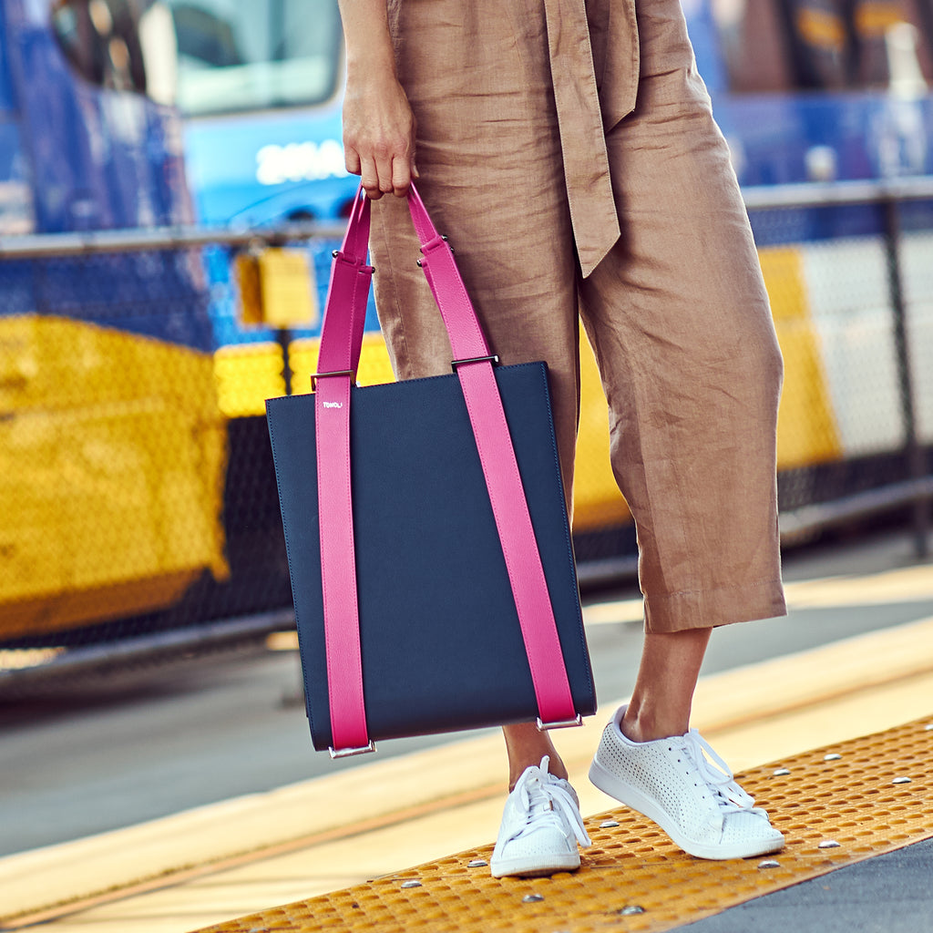 A fashion and style editorial photo of a woman holding a dark blue leather tote bag. The bag has a rectangular shape with pink straps. This is the Tomoli Kora Convertible Leather Tote in Flirty Denim.