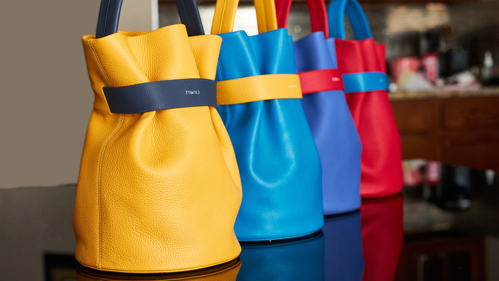 Colorful leather handbags in a perspective view. These are the La Bourse belted bucket bags from the Tomoli brand. They have a round base and contrasting belt cinch and top handle.