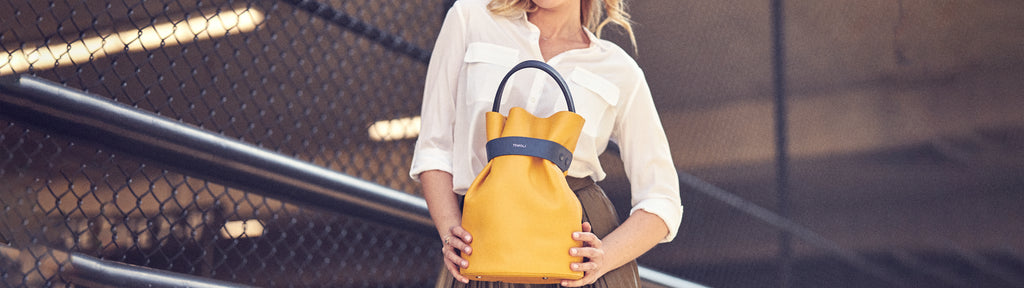 An editorial photo showing a model wearing a neutral Spring outfit and holding a yellow leather bucket bag. The bag has a top handle and a contrasting dark blue belt cinch. This is the Tomoli La Bourse bucket bag in Shady Maize.