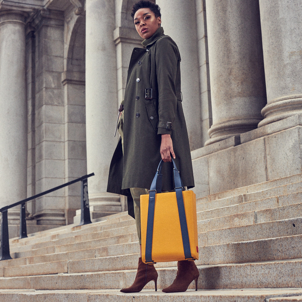 An outfit and style inspiration photo on how to wear a large tote bag. A woman wearing a green trench coat is holding a large yellow leather tote handbag. The bag has a structured rectangular briefcase shape and is color-blocked with dark blue straps. This is the Tomoli Kora large convertible leather tote in Shady Maize.