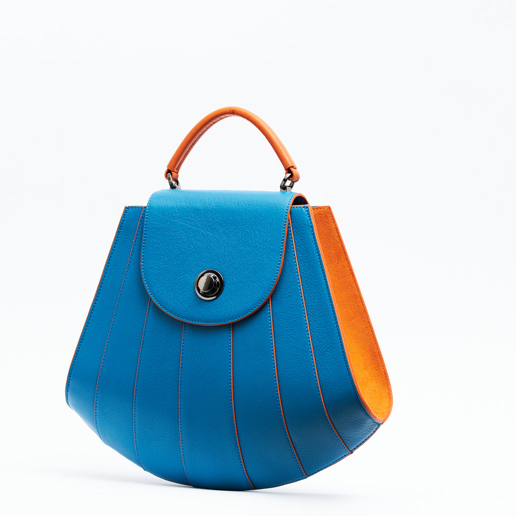 The product photo showing the side perspective of a colorful orange and blue leather handbag. The bag has a tapered trapeze shape that looks like a seashell. The bottom of the bag is rounded and there are orange radial lines and orange suede sides that create a color-blocked look. This is the Tomoli Gisel tapered top handle handbag in Blazing Sky.