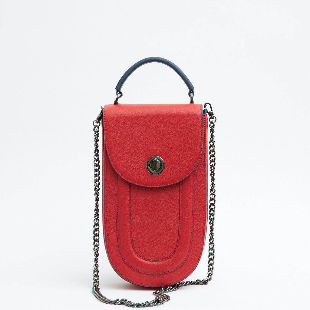A fashion product photo showing the front view of a colorful red leather handbag. The bag has an elongated saddle shape with a flap closure, a dark blue top handle, and dark blue trims. There is a chain strap and a metal lock in gunmetal hardware. This is the Tomoli Fitini II structured saddle handbag in Shady Red. This handbag can be used as an everyday crossbody bag, a work bag, or a special occasion bag.