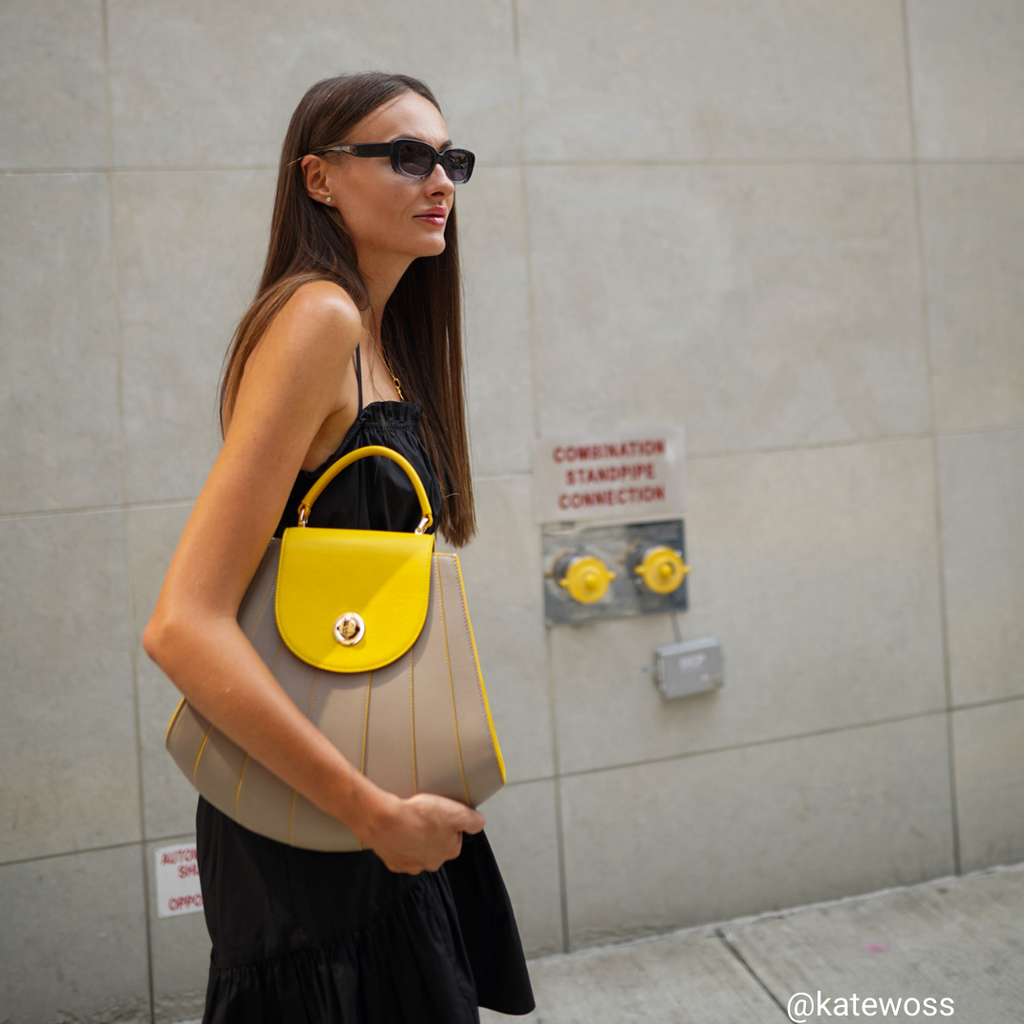 A fashion and style editorial photo showing influencer @katewoss wearing the Tomoli Gisel handbag in Sun Stone. The bag is color-blocked with yellow and beige and has a shell-like shape with a rounded flap closure.