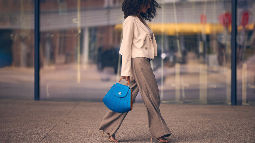 A fashion editorial photo showing a woman wearing a neutral outfit and holding a colorful blue top handle leather handbag. This is the Tomoli Gisel bag in Blazing Sky.