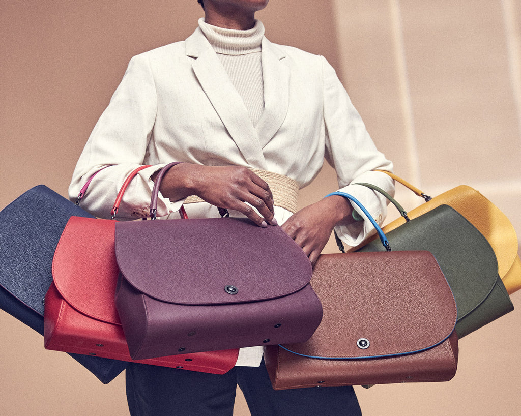 A fashion editorial photo showing a model holding multiple leather flap handbags on her arms. The bags are brown, yellow, green, red, blue, and dark purple. 