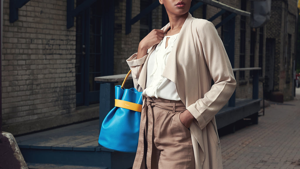 A fashion and style editorial photo showing a woman wearing a neutral outfit and holding a colorful leather bucket handbag. The bag is the Tomoli La Bourse bucket bag in Golden Sky.