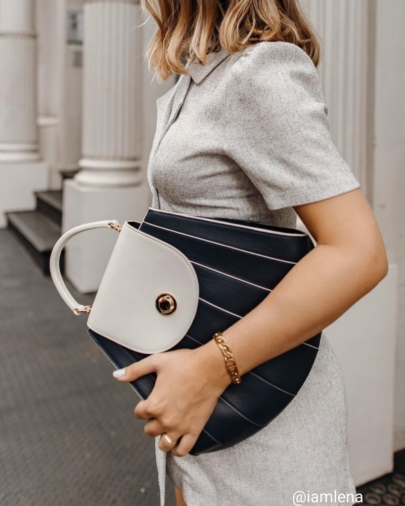 A woman's outfit photo showing influencer iamlena wearing a neutral outfit with a dark blue leather handbag. The bag has an off-white flap closure and thin stripes. This is the Tomoli Gisel bag in Ivorian Denim. It's the perfect work bag and everyday bag.