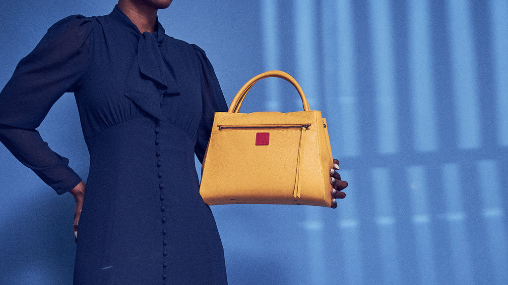 A maize yellow leather handbag held by a model on a blue background. The bag is the Briffani style by the brand Tomoli. It has a trapeze shape and top handles.