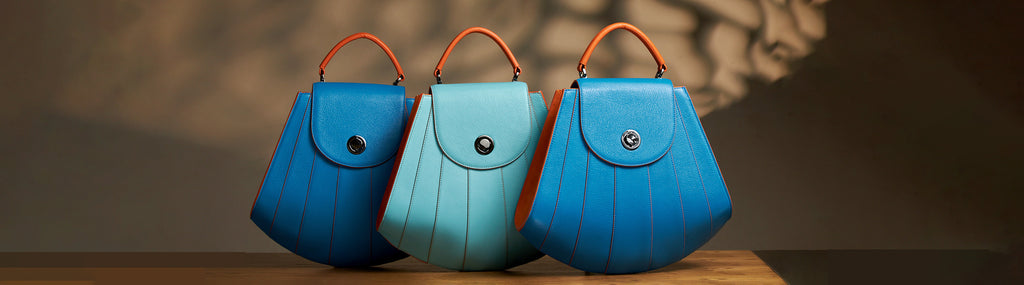 A fashion editorial photo showing 3 blue leather handbags. The handbags have a shell-like shape and radial contrasting lines. The orange handles create a color-blocking effect. These are the Tomoli Gisel handbags in aqua blue.