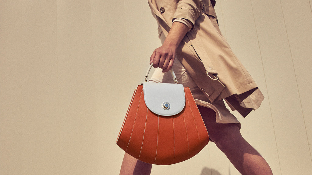 A fashion and style photo showing a model holding a color-blocked leather handbag. The bag has an orange body with an ivory flap closure and top handle. The shape of the bag resembles a seashell, with radial line detailing. This is the Tomoli Gisel handbag in Ivorian Blaze.