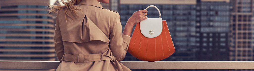 A fashion and style editorial photo showing a woman holding an orange leather work handbag. The handbag has a tapered silhouette and off-white contrasting line details. This is the Tomoli Gisel top handle handbag in Ivorian Blaze.