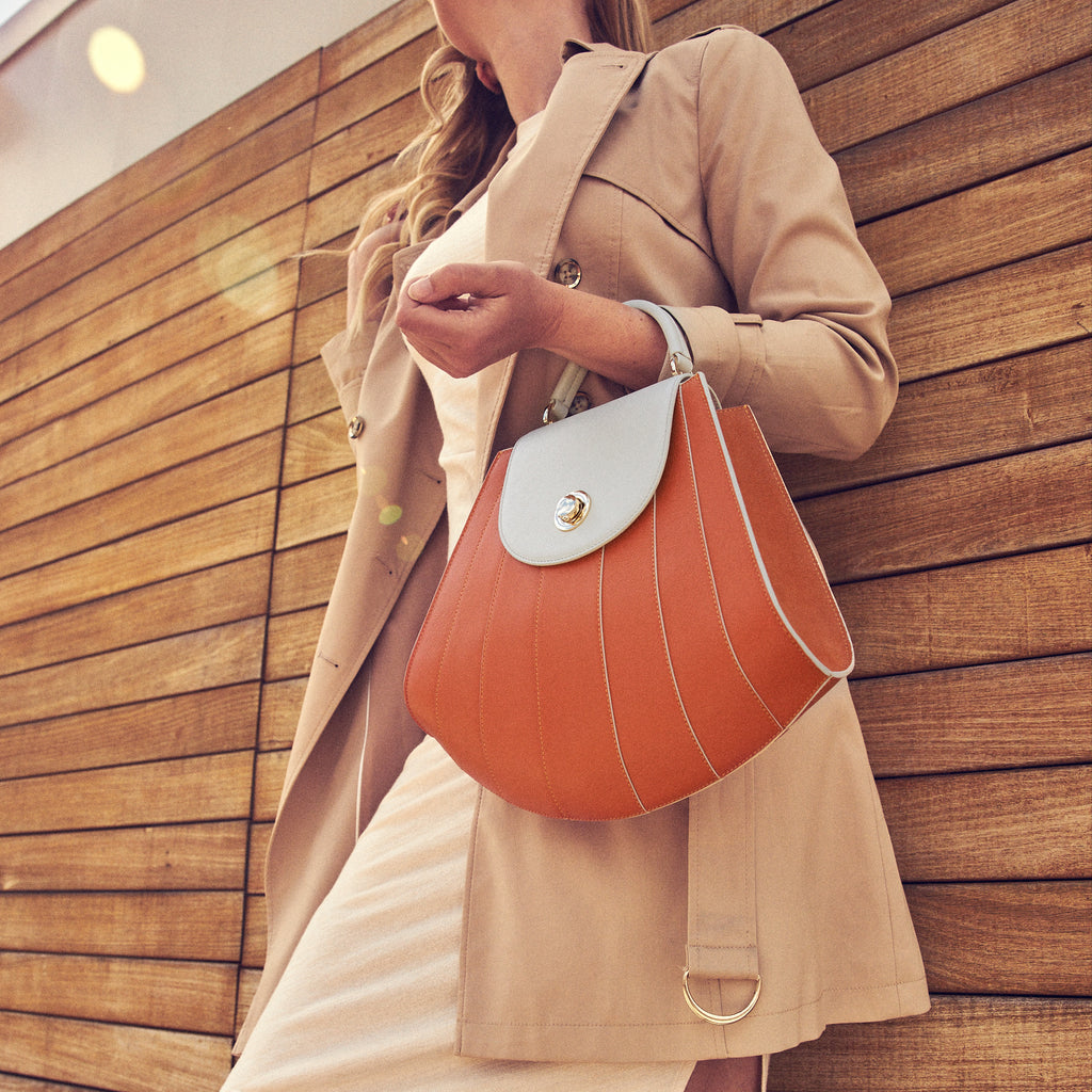 A fashion and style editorial photo showing a woman holding an orange leather handbag. The handbag has a tapered silhouette and off-white contrasting line details. This is the Tomoli Gisel top handle handbag in Ivorian Blaze.