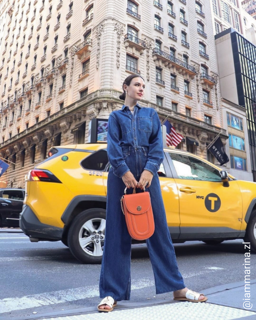 A fashion and style photo showing an influencer wearing a denim outfit and holding an orange leather handbag. The bag has a rounded shape and a flap closure with a top handle. This is a street style photo in New York City with a yellow cab.