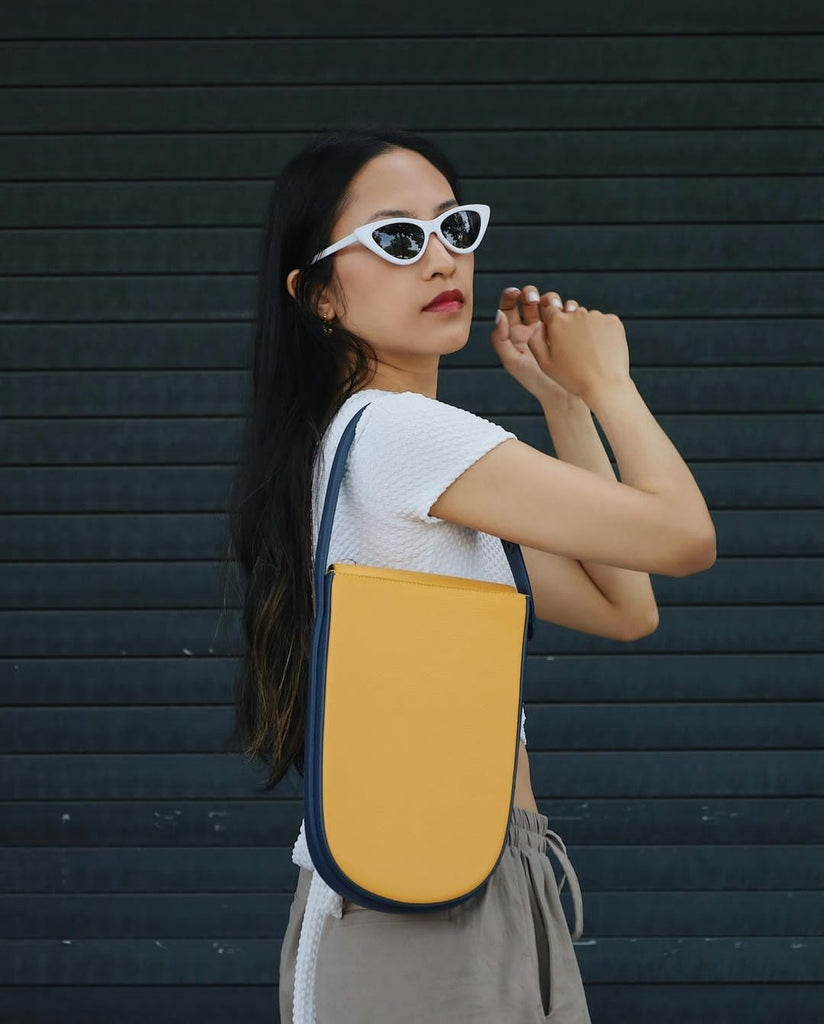 A fashion and style editorial photo showing influencer @cardiganfordays wearing white sunglasses and a colorful yellow handbag. The bag has a rounded shape and is color-blocked with a dark blue contouring handle.