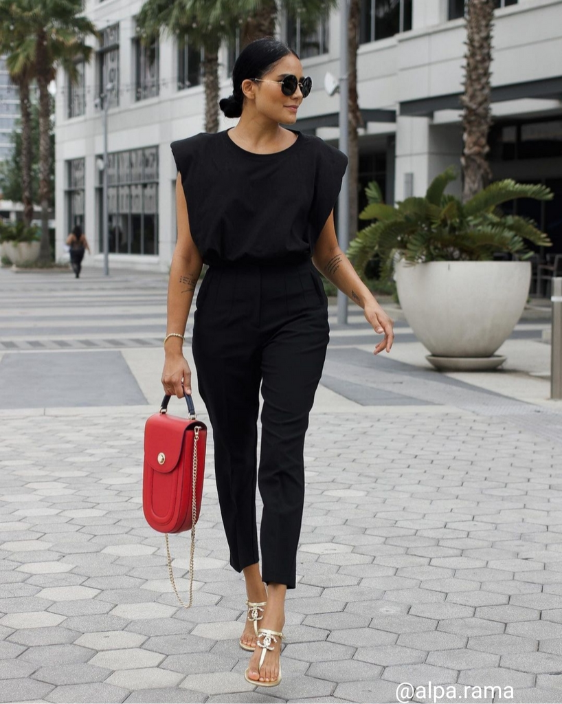 A fashion and style editorial photo showing influencer Alpa Rama wearing a black outfit and holding a red leather handbag. The handbag has an oblong silhouette with a flap closure and a top handle. The chain strap is hanging from the sides. This is the Tomoli Fitini II in Shady Red.