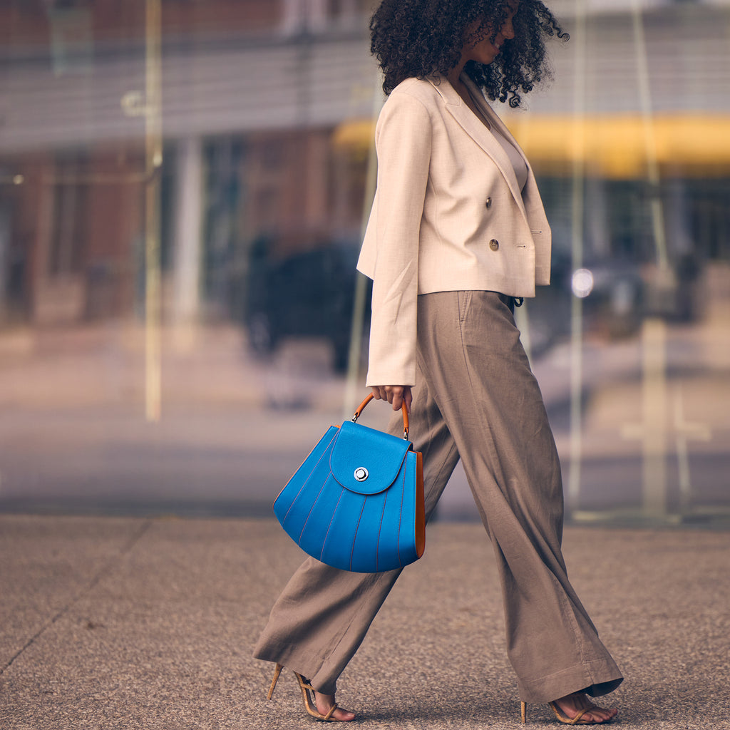 A fashion editorial photo showing a model holding a luxury leather handbag. The handbag has a shell-like shape and radial contrasting lines. The orange handle creates a color-blocking effect. The Tomoli Gisel handbag in aqua blue.