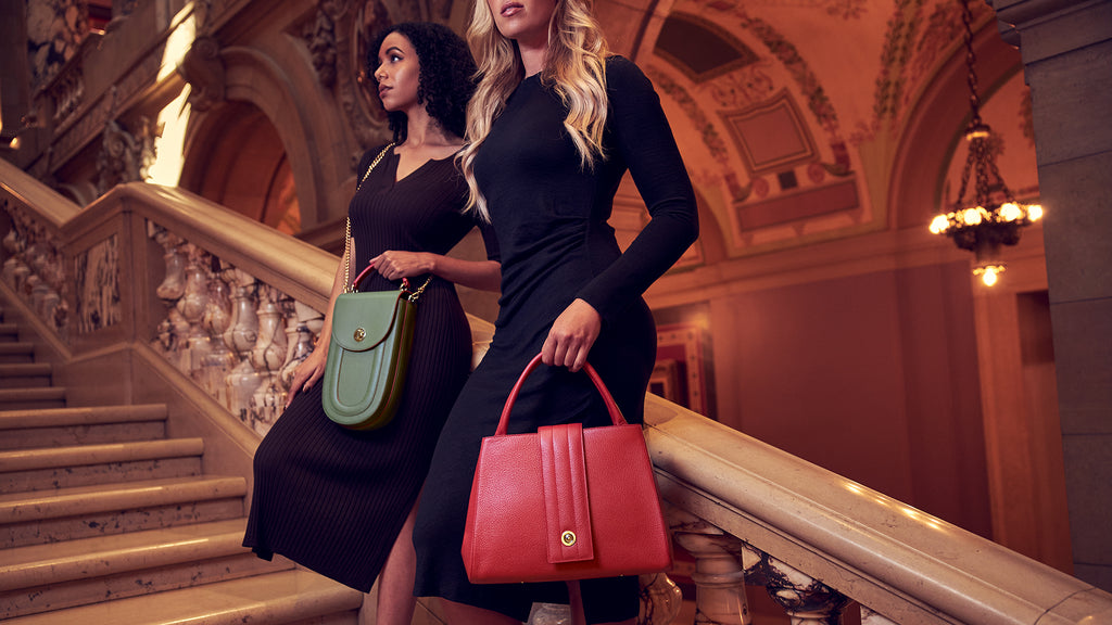 A fashion and style editorial photo showing two models holding colorful leather handbags. One has an oblong green bag and the other has a trapeze red tote bag. These are Tomoli's Fitini and Briffani handbags.