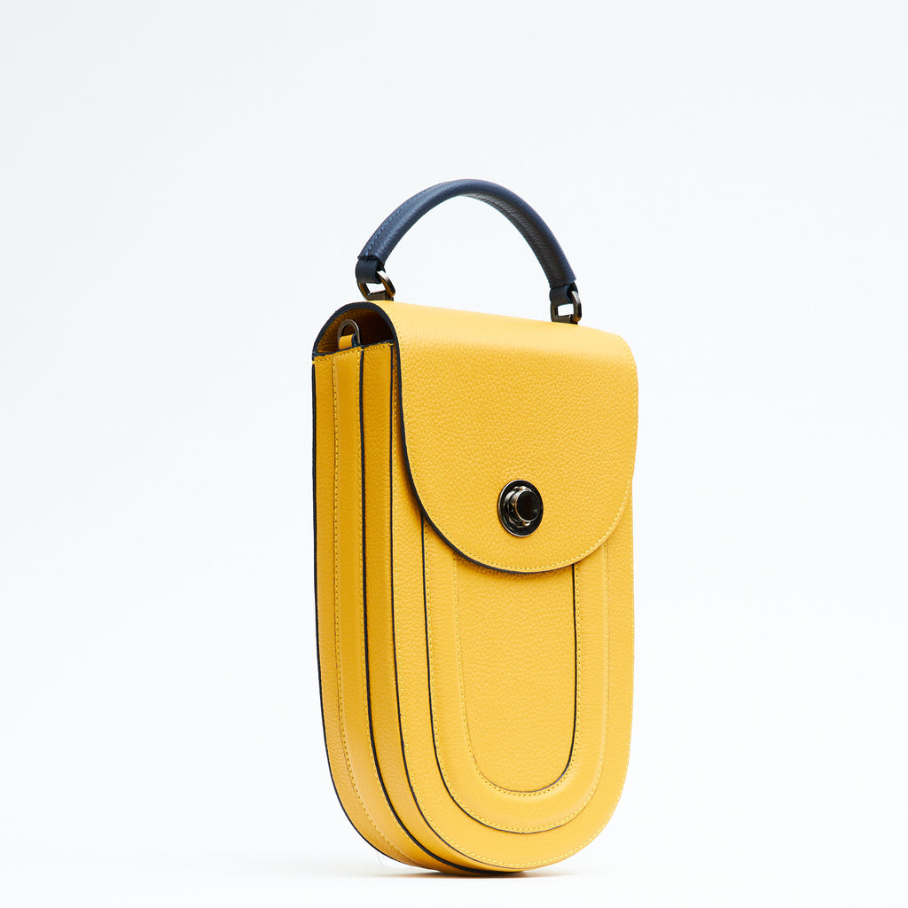 A fashion product photo showing the side  view of a colorful yellow leather handbag. The bag has an elongated rounded shape that resembles a saddle bag. The handbag has a flap closure with dark blue trims and a dark blue handle. There is a chain strap and a metal lock in gunmetal hardware. This is the Tomoli Fitini II structured saddle handbag in Shady Maize. This handbag can be used as an everyday crossbody bag, a work bag, or a special occasion bag.