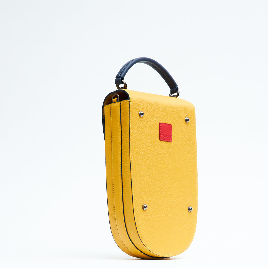 A fashion product photo showing the back view of a colorful yellow leather handbag. The bag has an elongated rounded shape that resembles a saddle bag. The handbag has dark blue trims and a dark blue handle. There is a small red square and gunmetal metal rivets.  This is the Tomoli Fitini II structured saddle handbag in Shady Maize. This handbag can be used as an everyday crossbody bag, a work bag, or a special occasion bag.