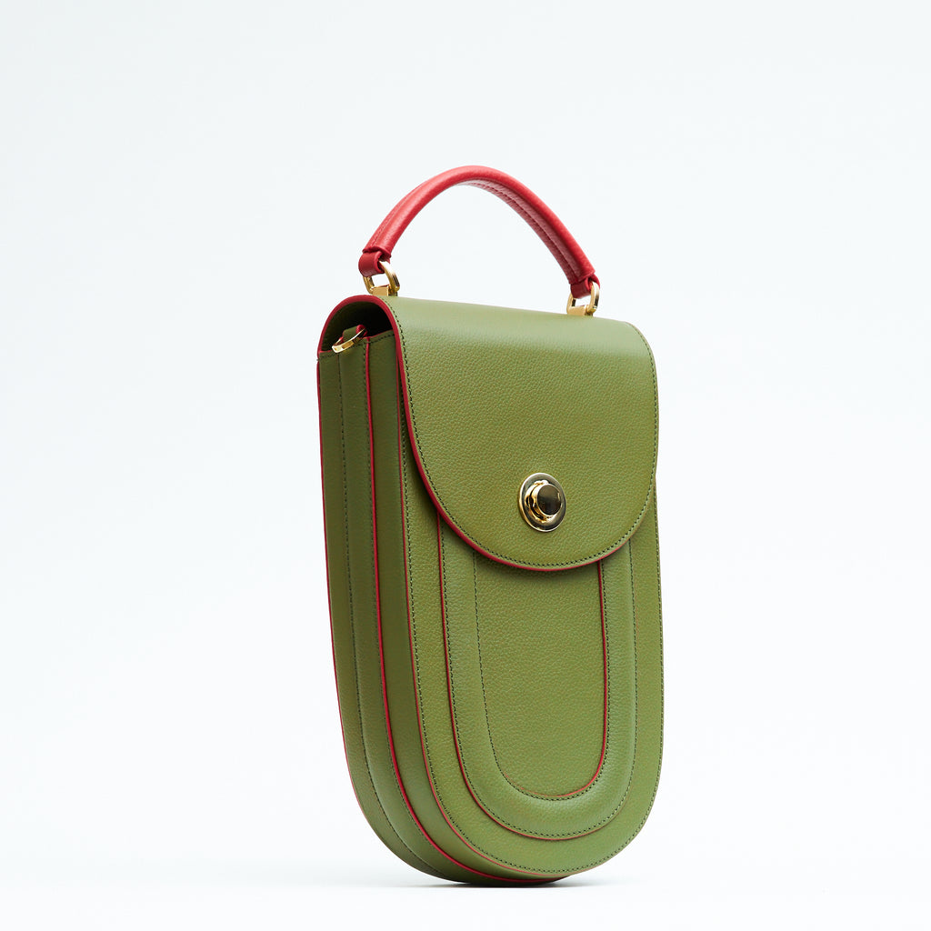 A fashion product photo showing the side view of a colorful green leather handbag. The bag has an elongated saddle shape with a flap closure, contrasting red trims, and a red handle. There is a metal lock in gold hardware. This is the Tomoli Fitini II structured saddle handbag in Hot Olive. This handbag can be used as an everyday crossbody bag, a work bag, or a special occasion bag.