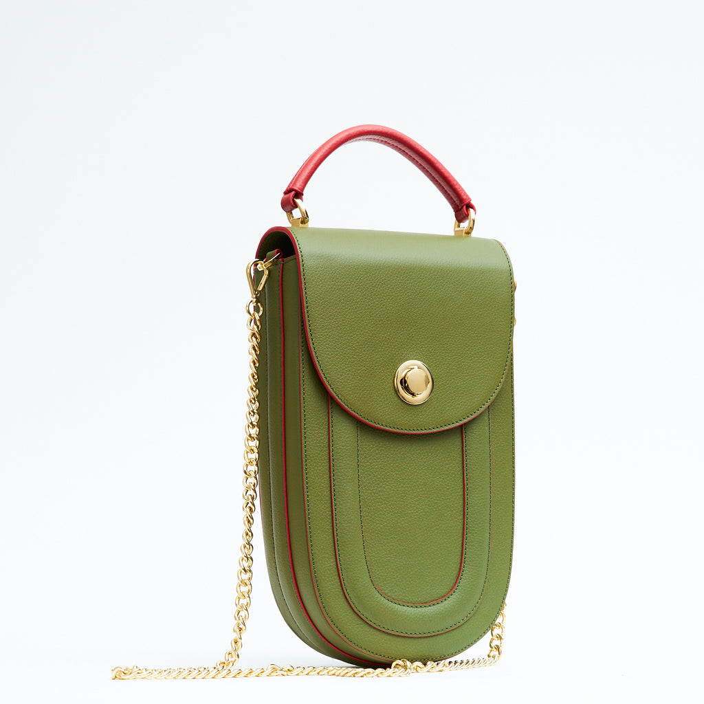 A fashion product photo showing the side view of a colorful green leather handbag. The bag has an elongated saddle shape with a flap closure, contrasting red trims, and a red top handle. There is a chain strap and a metal lock in gold hardware. This is the Tomoli Fitini II structured saddle handbag in Hot Olive. This handbag can be used as an everyday crossbody bag, a work bag, or a special occasion bag.