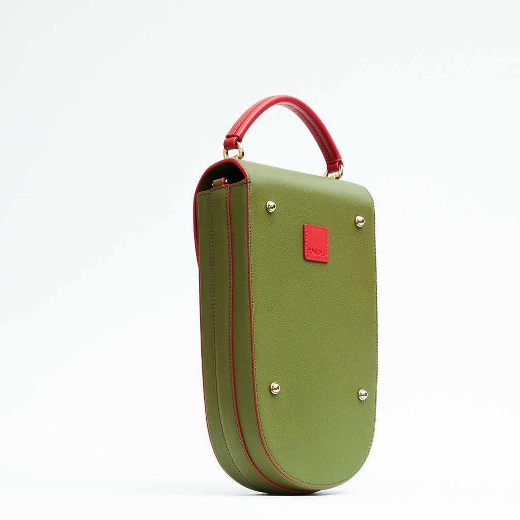 A fashion product photo showing the back view of a colorful green leather handbag. The bag has an elongated saddle shape with red trims and a red handle. There is a small red square and gold metal rivets. This is the Tomoli Fitini II structured saddle handbag in Hot Olive. This handbag can be used as an everyday crossbody bag, a work bag, or a special occasion bag.