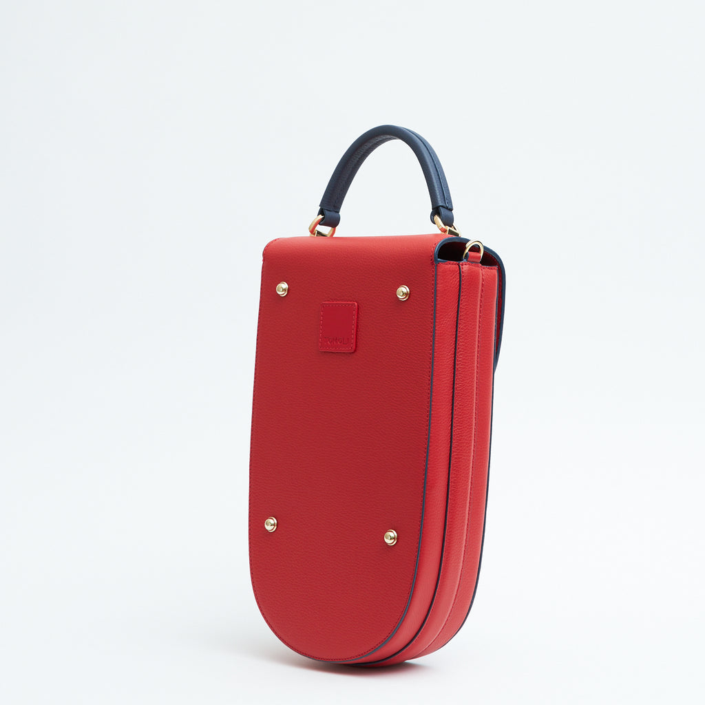 A fashion product photo showing the back view of a colorful red leather handbag. The bag has an elongated saddle shape with a dark blue top handle, and dark blue trims. There is a small red square and gold metal rivets. This is the Tomoli Fitini II structured saddle handbag in Shady Red. This handbag can be used as an everyday crossbody bag, a work bag, or a special occasion bag.