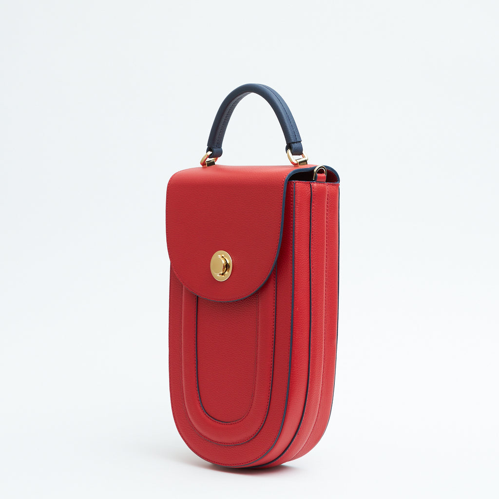 A fashion product photo showing the side view of a colorful red leather handbag. The bag has an elongated saddle shape with a flap closure, a dark blue top handle, and dark blue trims. There is a chain strap and a metal lock in gold hardware. This is the Tomoli Fitini II structured saddle handbag in Shady Red. This handbag can be used as an everyday crossbody bag, a work bag, or a special occasion bag.