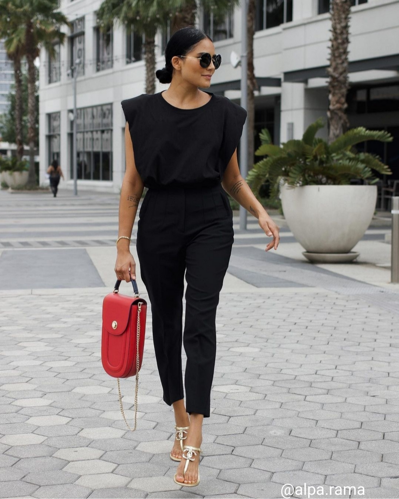 A fashion and style editorial photo showing influencer Alpa Rama wearing a black outfit with Tomoli's red Fitini leather handbag. The bag has a rounded saddle shape and a chain strap.