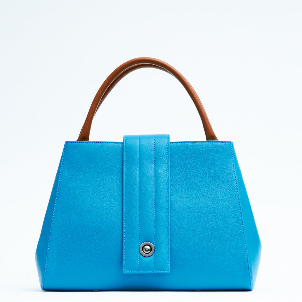 A product photo showing the front view of a colorful turquoise blue leather tote handbag. The handbag has a classic trapeze shape with brown top handles and a straight quilted flap in the middle. This is the Tomoli Briffani Lean interchangeable tote handbag in Brunette Sky. This bag can be used as an everyday bag, a work tote, or even an oversized clutch.