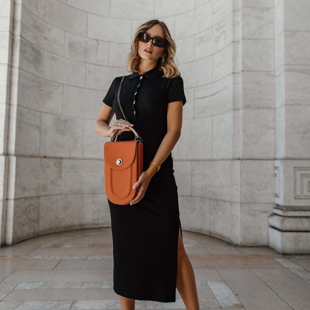 A fashion editorial photo showing a woman wearing a black dress and holding an orange leather bag. The bag has an elongated saddle shape, a contrasting off-white handle, and a chain strap. The overall look has a color-blocked effect with Halloween black and orange colors.