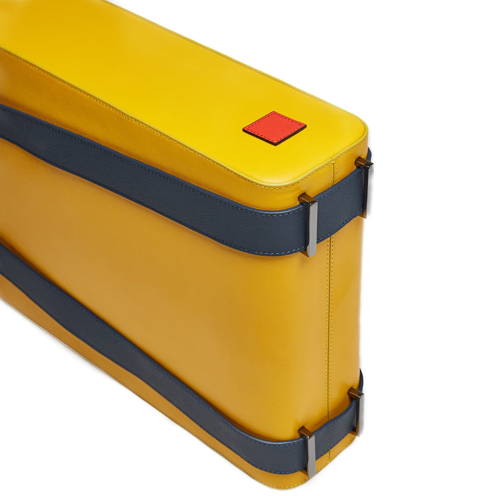 The product photo of a yellow leather tote bag. The tote bag has a structured rectangular briefcase shape and is color-blocked with dark blue leather straps. This is the bottom view of the Tomoli Kora large convertible leather tote in shady maize. The bag can fit a laptop and can be styled for work or to elevate casual outfits.