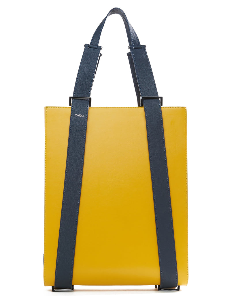The product photo of a yellow leather tote bag. The tote bag has a structured rectangular briefcase shape and is color-blocked with dark blue leather straps. This is the front view of the Tomoli Kora large convertible leather tote in shady maize. The bag can fit a laptop and can be styled for work or to elevate casual outfits.