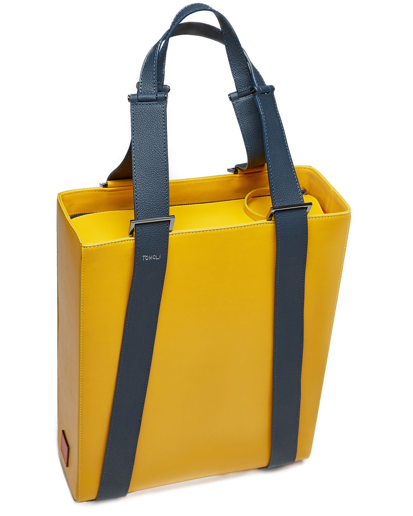 The product photo of a yellow leather tote bag. The tote bag has a structured rectangular briefcase shape and is color-blocked with dark blue leather straps. This is the side view of the Tomoli Kora large convertible leather tote in shady maize. The bag can fit a laptop and can be styled for work or to elevate casual outfits.
