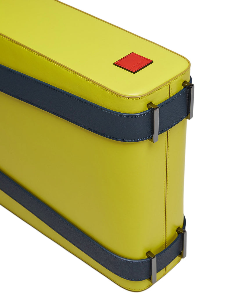 A product photo of a lime green leather tote bag. The tote bag has a structured rectangular briefcase shape and color-blocked dark blue leather straps. This is the bottom view of the Tomoli Kora large convertible leather tote in shady chartreuse. The bag can fit a laptop and can be styled for work or to elevate casual outfits.