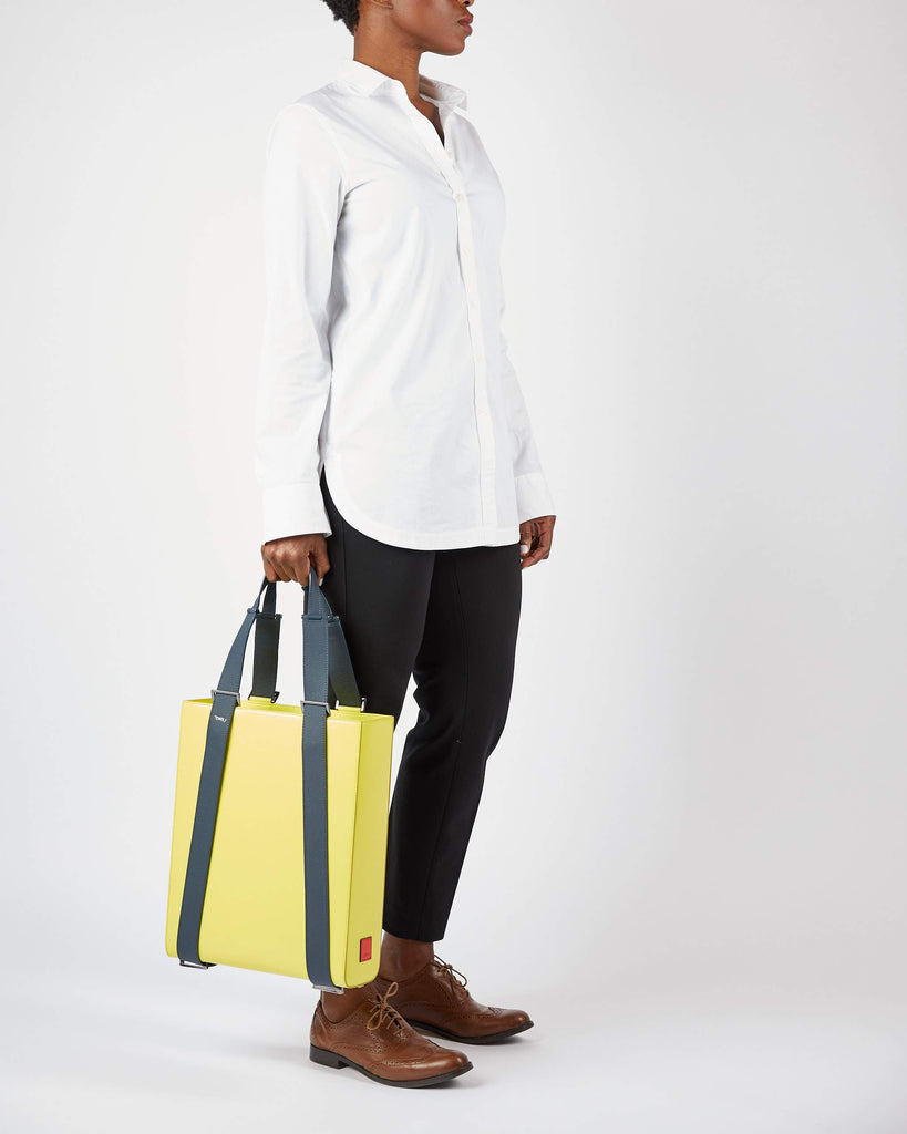 A fashion and style editorial photo showing a woman holding a lime green leather tote bag. The tote bag has a structured rectangular briefcase shape and color-blocked dark blue leather straps. This is the Tomoli Kora large convertible leather tote in shady chartreuse. The bag can fit a laptop and can be styled for work or to elevate casual outfits.