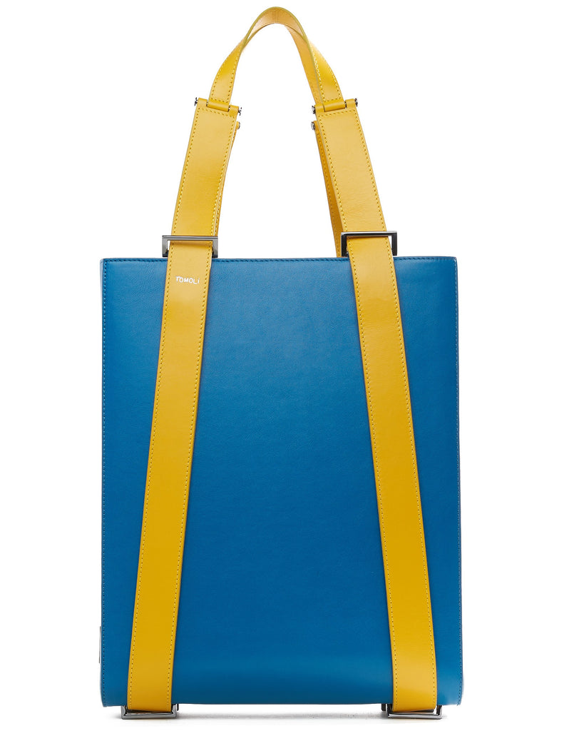 The product photo of a teal leather tote bag. The tote bag has a structured rectangular briefcase shape and color-blocked yellow leather straps. This is the front view of the Tomoli Kora large convertible leather tote in golden teal. The bag can fit a laptop and can be styled for work or to elevate casual outfits.