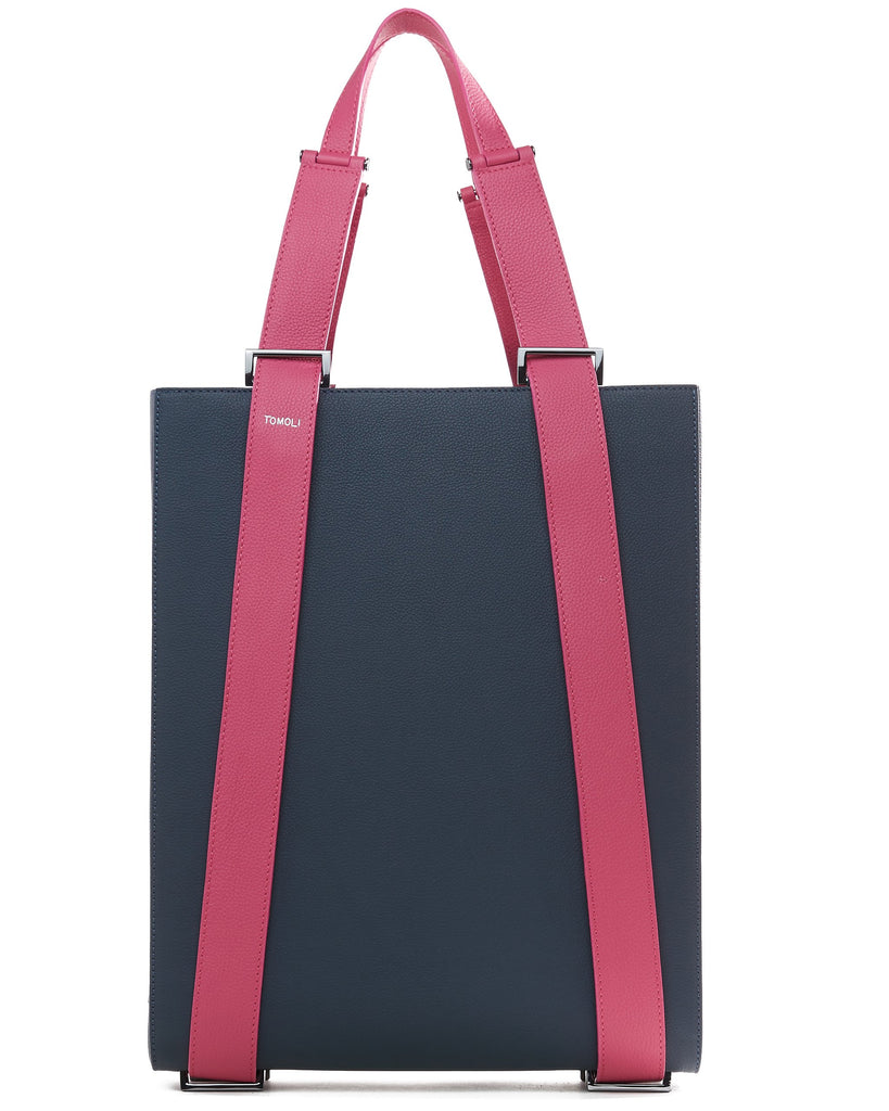 The product photo of a dark blue leather tote bag. The tote bag has a structured rectangular briefcase shape and color-blocked pink leather straps. This is the front view of the Tomoli Kora large convertible leather tote in flirty denim. The bag can fit a laptop and can be styled for work or to elevate casual outfits.