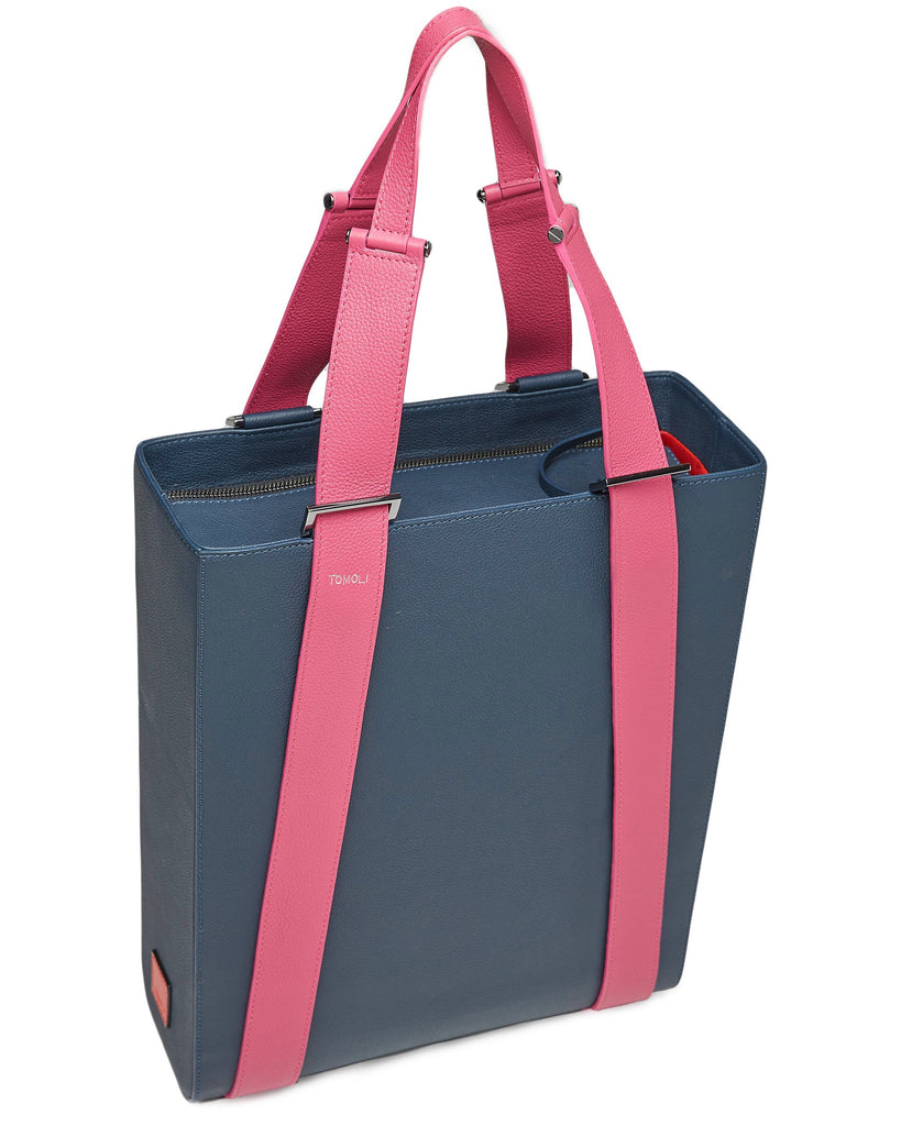 The product photo of a dark blue leather tote bag. The tote bag has a structured rectangular briefcase shape and color-blocked pink leather straps. This is the side view of the Tomoli Kora large convertible leather tote in flirty denim. The bag can fit a laptop and can be styled for work or to elevate casual outfits.