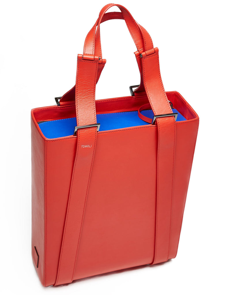 A product photo of a red leather tote handbag. The tote has a structured rectangular shape and flat handles. This is the Tomoli Kora tote in red aria leather. This bag can be used as a work bag or an everyday bag.