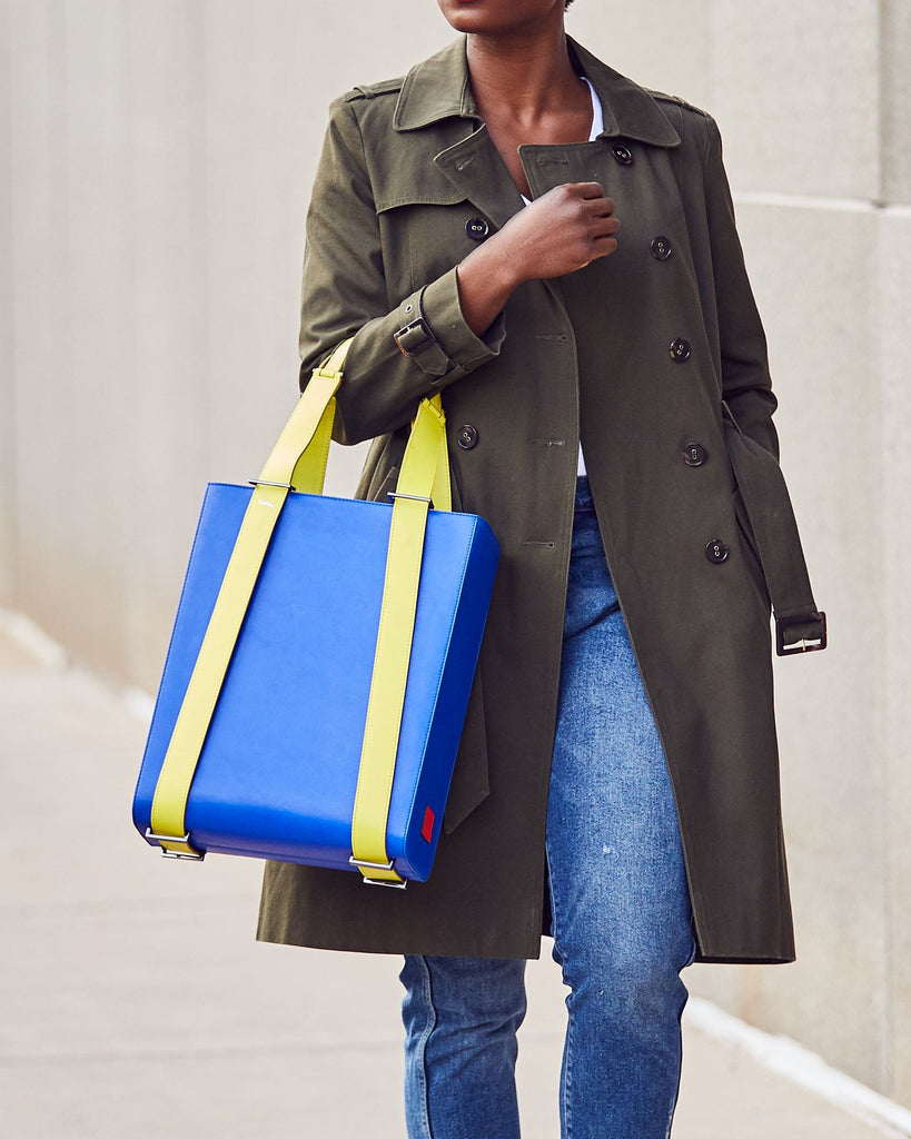 A fashion and style editorial photo showing a woman wearing a trench coat and holding a bright blue leather tote bag. The tote bag has a structured rectangular briefcase shape and color-blocked neon green leather straps. This is the Tomoli Kora large convertible leather tote in lime blue. The bag can fit a laptop and can be styled for work or to elevate casual outfits.