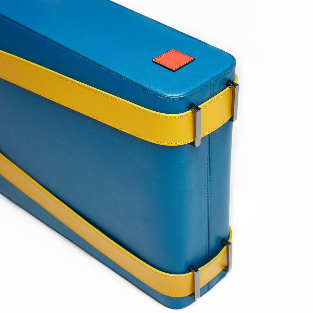 The product photo of a teal leather tote bag. The tote bag has a structured rectangular briefcase shape and color-blocked yellow leather straps. This is the bottom view of the Tomoli Kora large convertible leather tote in golden teal. The bag can fit a laptop and can be styled for work or to elevate casual outfits.