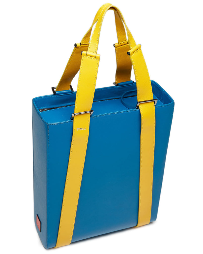The product photo of a teal leather tote bag. The tote bag has a structured rectangular briefcase shape and color-blocked yellow leather straps. This is the top view of the Tomoli Kora large convertible leather tote in golden teal. The bag can fit a laptop and can be styled for work or to elevate casual outfits.