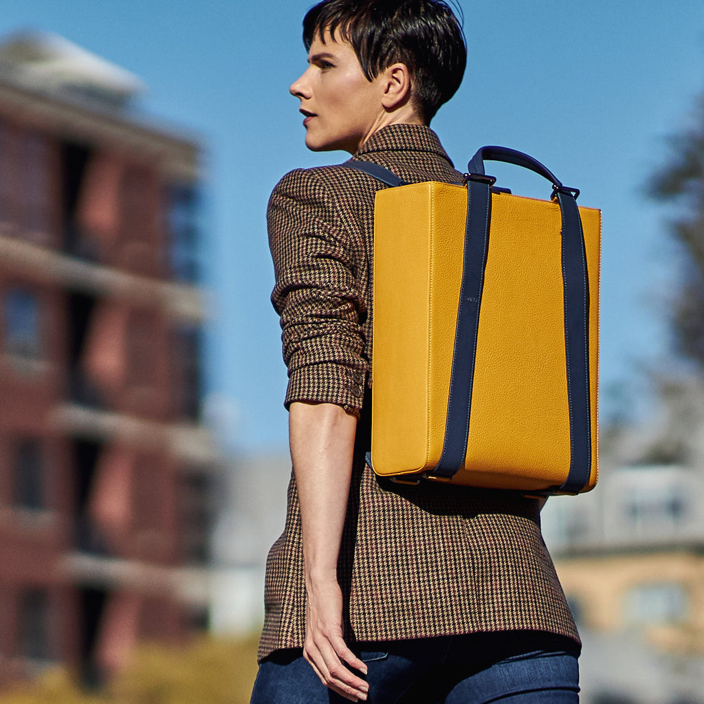 An outfit and style inspiration photo on how to wear a large tote bag. A woman wearing a neutral brown  blazer is carrying a large yellow leather tote handbag as a backpack. The bag has a structured rectangular briefcase shape and is color-blocked with dark blue straps. This is the Tomoli Kora large convertible leather tote in Shady Maize.