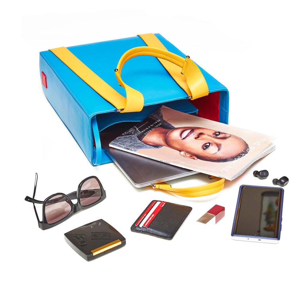 A fashion flat lay showing a turquoise blue handbag with yellow straps. The bag contains a magazine, a laptop, sunglasses, a phone, and a makeup case. This is the Tomoli Kora large convertible leather tote in Golden Sky.