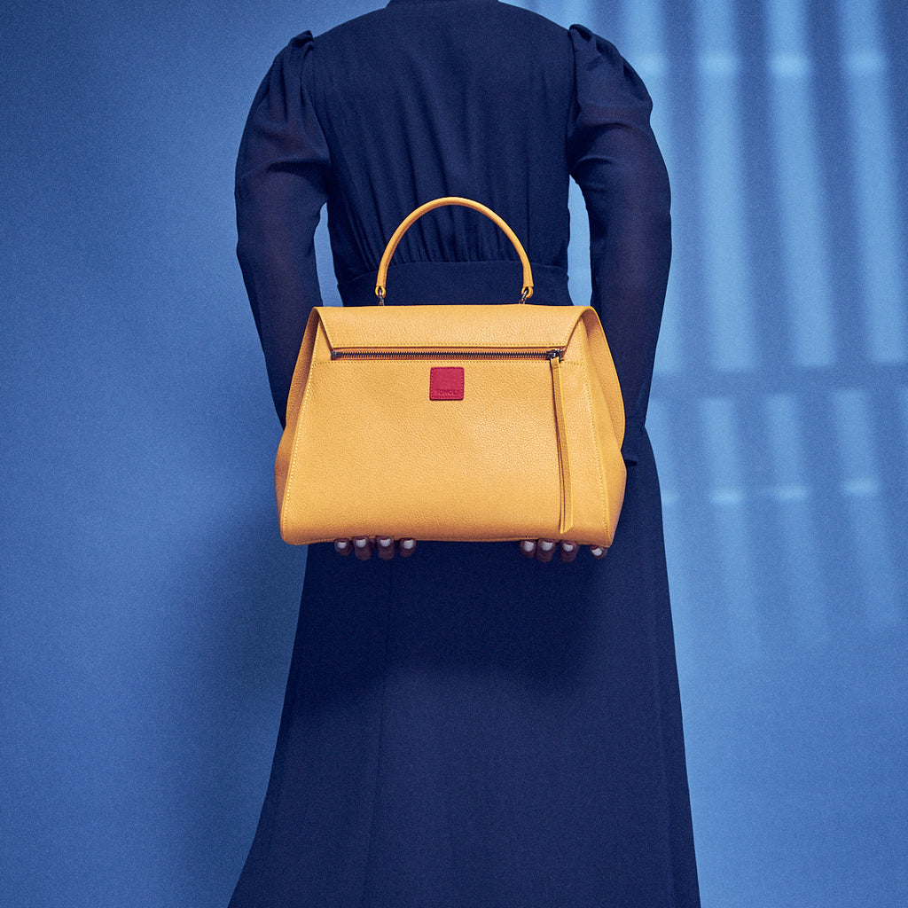 A fashion and style editorial photo showing a model holding a mustard yellow top handle leather handbag. The back of the handbag has a zipper attachment and a small red square. The yellow bag creates a color-blocking effect against the model's blue dress. This is the Tomoli Briffani Jut interchangeable satchel in Maize.