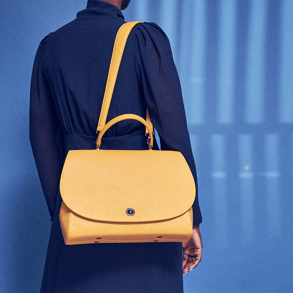 A fashion and style editorial photo showing a model wearing a mustard yellow flap leather handbag. The handbag has an oversized round flap cover and a wide shoulder strap. The yellow bag creates a color-blocking effect against the model's blue dress. This is the Tomoli Briffani Jut in Maize.