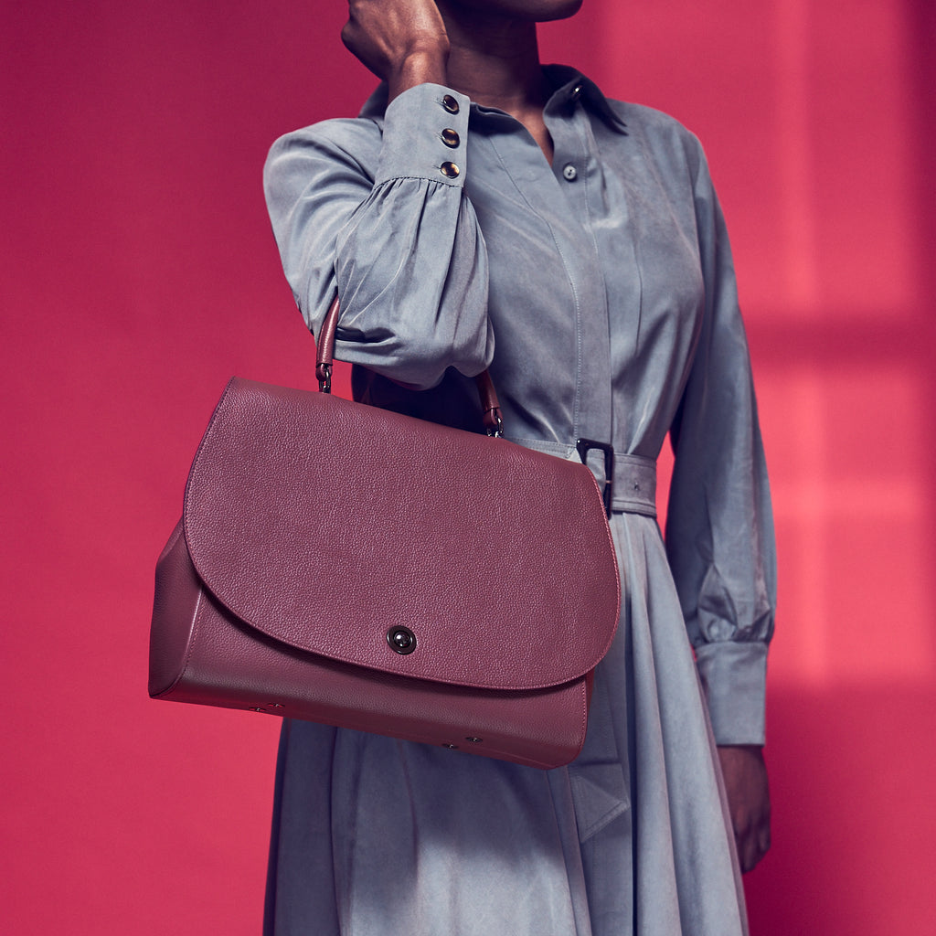 A fashion and style photo showing a model holding a burgundy red flap leather handbag. The handbag has a minimalistic design and an oversized round flap cover. The bag creates a color-blocking effect against the model's light blue dress. This is the Tomoli Briffani Jut interchangeable satchel in Sangria.