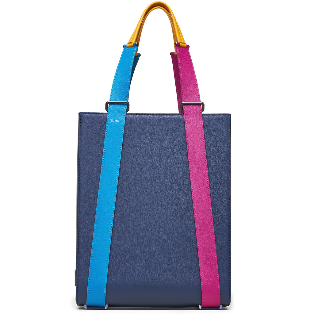 The product photo of a dark blue leather tote bag. The tote bag has a structured rectangular briefcase shape and color-blocked blue and pink leather straps. This is the front view of the Tomoli Kora large convertible leather tote in eclectic denim. The bag can fit a laptop and can be styled for work or to elevate casual outfits.