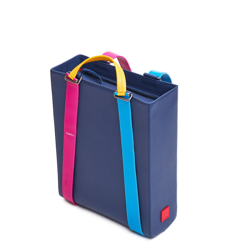 The product photo of a dark blue leather tote bag. The tote bag has a structured rectangular briefcase shape and color-blocked blue and pink leather straps. This is the side view of the Tomoli Kora large convertible leather tote in eclectic denim. The bag can fit a laptop and can be styled for work or to elevate casual outfits.